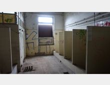 Ein verlassenes und heruntergekommenes Badezimmer mit gelblichen Fliesen, mehreren offenen Kabinen und Graffiti an den Wänden. Ein einzelnes, beschädigtes Fenster lässt Licht in den Raum, während die Wände und der Boden deutliche Spuren von Vernachlässigung zeigen.