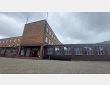 Backstein-Gebäude mit flachem Haupttrakt und einem leicht erhöhten seitlichen Turmbereich, großen Fenstern mit roten Rahmen und einem gepflasterten Vorplatz. Der Eingangsbereich ist mit Glasbausteinen und einer Doppeltür gestaltet.