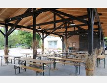 Überdachter Außenbereich mit Holzbänken und -tischen, dunklen Holzstützen und einem Holzdach; dekoriert mit gebundenem Getreide. Im Hintergrund befinden sich Bäume, ein Weg und ein Gebäude.