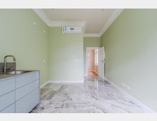 Leerer, schmaler Küchen- oder Hauswirtschaftsraum mit hellgrünen Wänden, glänzendem Marmorfliesenboden und einer kleinen Küchenzeile mit Spüle links. An der Stirnwand ist eine Klimaanlage montiert; eine offene Tür führt in einen angrenzenden Flur mit Fensterlicht.