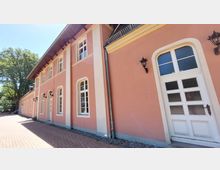 Rosa Gebäude mit weißen Fensterrahmen und Türen, ergänzt durch dekorative Außenleuchten. Das Gebäude befindet sich an einem gepflasterten Weg und ist von grünen Bäumen umgeben.