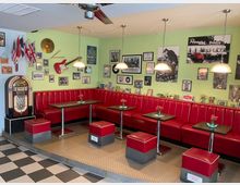 Innenraum eines Retro-Diners mit langen roten Kunstleder-Sitzbänken, kleinen quadratischen Tischen und passenden Hockern auf einem schwarz-weiß karierten Boden. An der hellgrünen Wand hängen zahlreiche gerahmte Fotos und Musikdekorationen, daneben steht eine Jukebox; mehrere Hängelampen beleuchten die Sitzreihe.