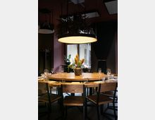 Ein gemütlicher Innenraum mit einem runden Holztisch, umgeben von Holzstühlen. Der Tisch ist mit Gläsern, Besteck und einer Vase mit gelben Blumen dekoriert, während eine große, hängende Lampe mit Pflanzen darüber hängt; im Hintergrund befindet sich ein Fenster und ein dunkler Vorhang.