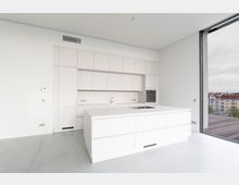 Moderne, minimalistische Küche mit weißen Einbauschränken, einer Kücheninsel mit integriertem Kochfeld und Spüle sowie einem großen, bodentiefen Fenster mit Blick auf ein Stadtviertel mit mehrstöckigen Gebäuden.