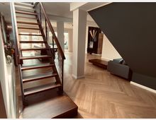 Moderne Wohnraumgestaltung mit offenem Grundriss, einer Holztreppe mit Metallgeländer, hellem Fischgrätenparkettboden, großer Fensterfront im Hintergrund und einer Sitzecke mit Sofa. Links steht eine Vase mit Blumen auf einem seitlichen Abstellbereich.