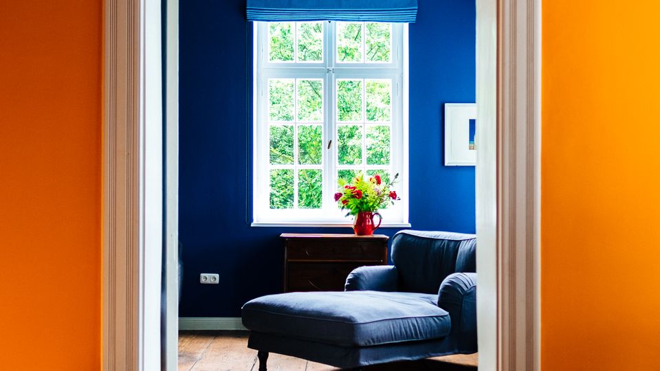 Blick durch einen weißen Türrahmen aus einem Raum mit orangefarbenen Wänden in ein Wohnzimmer mit dunkelblauer Wand, großem Sprossenfenster und blauem Raffrollo. Vor dem Fenster stehen ein dunkler Sessel mit Ottomane, ein kleiner Tisch mit Vase sowie ein gemusterter Teppich auf Holzboden.