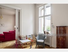 Wohnzimmer mit hohen Decken und Holzfußboden in einem Altbau. Im Vordergrund stehen Kinderstühle, ein Kindertisch und ein geflochtener Sessel vor einem großen Sprossenfenster, rechts ein antikes Holzsideboard, im angrenzenden Raum ein rotes Sofa.
