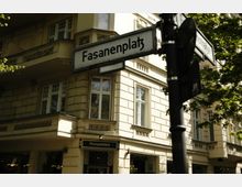 Straßenschild mit der Aufschrift „Fasanenplatz“ vor einem hellen, historischen Gebäude mit dekorativen Fassadenelementen, großen Fenstern und einem Geschäft für Antiquitäten im Erdgeschoss. Im Hintergrund sind teilweise Bäume und weitere Gebäude sichtbar.