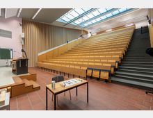 Ein Hörsaal mit ansteigenden Sitzreihen aus Holz und integrierten Klapptischen, einer Treppe auf der rechten Seite, einem Dozentenpult mit Mikrofon links und einem Tisch im Vordergrund. Das Glasdach sorgt für Tageslicht, während die Vertäfelung an der Wand schallabsorbierend wirkt.