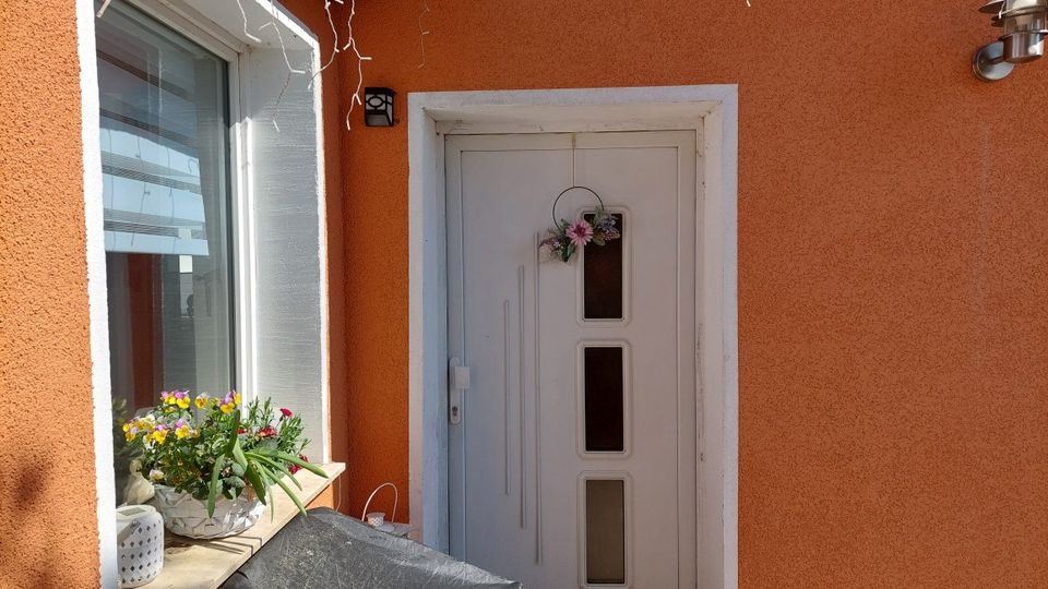 Eingangsbereich eines Wohnhauses mit orangefarbener Putzfassade, weißer Haustür mit drei schmalen Glasfeldern und kleinem Türkranz. Links daneben ein großes Fenster mit Blumenkasten auf der Fensterbank sowie eine Außenwandleuchte.