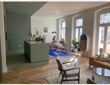 Offener Wohnraum mit integrierter Küche und Essbereich. Die grüne Küche mit Kochinsel grenzt an einen hellen Wohnbereich mit Holzboden, großen Fensterfronten, einem gelben Sofa, einem blauen Teppich und mehreren Pflanzen.