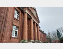 Backsteingebäude mit klassizistischer Architektur, gekennzeichnet durch hohe Säulen und ein dreieckiges Giebeldach mit Verzierungen. Im Umfeld sind Bäume und ein grauer, bewölkter Himmel sichtbar.