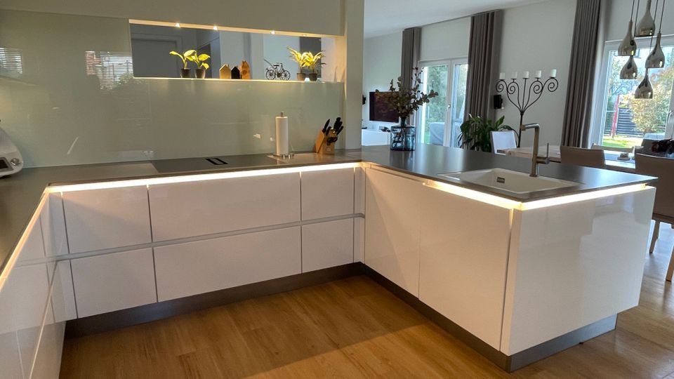 Moderne Küche mit glänzenden weißen Einbauschränken und einer Arbeitsplatte aus dunkelgrauem Material, beleuchtet durch integrierte LED-Leisten. Im Hintergrund befinden sich ein Essbereich mit Tisch und Stühlen sowie bodentiefe Fenster mit Vorhängen, die Blick auf einen Garten ermöglichen.