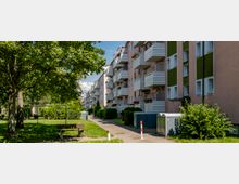 Mehrstöckige Wohngebäude mit Balkonen in einer Reihe, umgeben von gepflegten Grünflächen und Bäumen; ein Gehweg mit Sitzbank sowie kleine Büsche und Versorgungsboxen im Vordergrund.