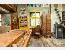 Innenraum einer Werkstatt mit rustikalem Holzboden. Zu sehen sind eine große Werkbank, ein gelbes Regal mit Glasfront, eine Schubladenkommode, ein Holzofen mit Rohr sowie Wandbilder und Geräte verteilt im Raum.