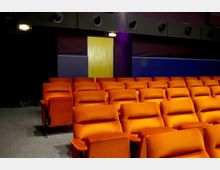 Kinosaal mit orangefarbenen Polstersitzen in mehreren Reihen, dunklen Wänden und einer gelben Tür im Hintergrund. Oberhalb der Wände befinden sich Lüftungsöffnungen, und der Boden ist mit Teppichboden bedeckt.