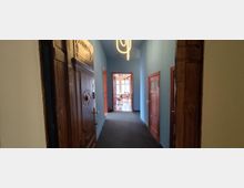 Langer Flur in einem Altbau mit blau gestrichenen Wänden, dunklem Teppichboden und großen, dekorativ geschnitzten Holztüren. An der Decke hängt eine moderne Leuchte, am Ende führt eine offene Tür in einen hellen Raum mit großen Fenstern.