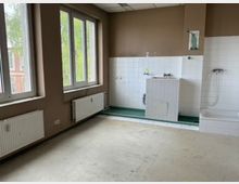 Leerer, teilweise gefliester Sanitärraum mit großen Fenstern, Heizkörpern und beigefarbenen Wänden. An der rechten Wand befinden sich ein gefliester Wasch-/Wasseranschlussbereich und eine Badewanne mit Duscharmatur; der Boden ist unbedeckt und wirkt unfertig.