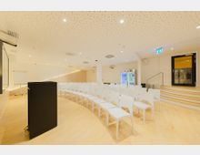 Moderner Seminarraum mit hellen Holzfußböden und weißen Wänden; Bestuhlung mit weißen Kunststoffstühlen in Reihen angeordnet, ein schwarzes Rednerpult vorne. Im Hintergrund ein Flipchart, ein Getränkeautomat und eine schwarze Tür mit Notausgangsschild.