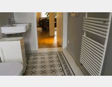 Badezimmer mit einer weißen Wand, einem Waschbecken mit Wasserhahn, einem Schrank sowie einem Handtuchheizkörper. Der Boden ist mit dekorativen Fliesen gestaltet, und eine geöffnete Tür führt zu einem angrenzenden Raum mit Holzboden und Möbeln.