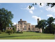 Schloss Babelsberg, ein historisches Schloss im neugotischen Stil mit hellen Fassade, Türmen und Zinnen, umgeben von einer weitläufigen Grünanlage mit Bäumen, unter blauem Himmel.
