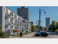 Straßenszene mit modernen Wohngebäuden links, weißer Fassade und Balkonen mit Blumen. Im Hintergrund sind ein hoher Büroturm mit der Aufschrift „Der Oderturm“ sowie Bäume und Straßenlaternen zu sehen, rechts verläuft eine Straßenkreuzung mit geparkten Autos und Straßenverkehr.