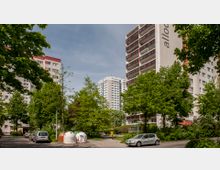 Mehrstöckige Wohngebäude in einer urbanen Umgebung, umgeben von grünen Bäumen und Sträuchern. Im Vordergrund befinden sich geparkte Autos und Recyclingcontainer auf einer kleinen Straße.