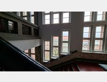 Helles Treppenhaus in einem mehrstöckigen Gebäude mit großen, rechteckigen Fensterfronten und Blick auf umliegende Backsteinbauten. Zu sehen sind ein Treppenlauf mit dunklem Geländer, geflieste Wandbereiche und ein offener Zwischenbereich mit Oberlichtfenstern.