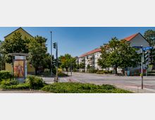 Straßenkreuzung in einem Wohngebiet mit mehrstöckigen Wohnhäusern mit roten Dächern, umgeben von Bäumen. Im Vordergrund steht eine Werbesäule, daneben Ampeln und Straßenschilder für die Potsdamer Straße und den Anton-Saefkow-Ring.