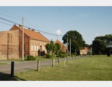 Backsteingebäude mit Satteldach und weißen Fensterrahmen in einer ländlichen Umgebung. Im Vordergrund führen ein Gehweg und eine Wiese, umgeben von Holzpfosten, entlang der Gebäude.