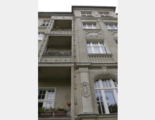 Fassadenansicht eines historischen Wohngebäudes mit verzierten Stuckelementen, großen Fenstern und Balkonen, die teilweise bepflanzt sind. Die Architektur zeigt typische dekorative Details wie Reliefs und Balustraden.