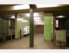 U-Bahn-Station mit grünen Wandfliesen, gelben Fahrkartenautomaten und einem Treppenaufgang im Hintergrund, der von Graffiti und Plakaten gesäumt ist. Metallpfeiler und Signalanlagen sind im Vordergrund zu sehen.