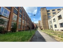 Industriegelände mit alten Backsteingebäuden und großflächigen Fenstern, flankiert von betonierten und weißen Gebäudefassaden. Ein schmaler Gehweg und Grünflächen befinden sich im Vordergrund, mit einem wolkigen Himmel im Hintergrund.