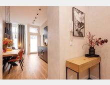 Heller, moderner Wohn- und Essbereich mit langem Holztisch und Lederstühlen, schwarzer Küchenzeile sowie hohen Fenstern und Glastür mit grauen Vorhängen. Im Vordergrund steht eine schmale Holz-Konsole mit Vase und Wandbild an einer weißen Trennwand; Holzfußboden und Deckenspots prägen den Raum.