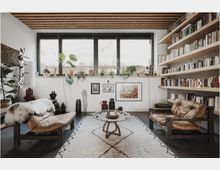 Wohnzimmer mit modernem Design: Zwei braune Ledersessel, ein runder Couchtisch und ein beige gemusterter Teppich. An einer Wand befinden sich große Fenster mit Pflanzen auf der Fensterbank, an der anderen Wand ein Regal mit zahlreichen Büchern.