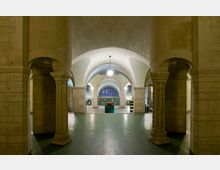 Großzügiger Innenraum mit mehreren Rundbögen und massiven Säulen aus Stein, die einen symmetrischen Durchgang bilden. Im Hintergrund zieren hohe Fenster die Wand, und eine zentrale rote Skulptur steht im Blickpunkt.