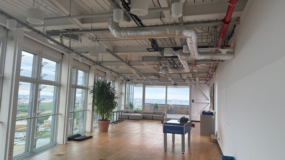 Open-plan modern office lounge with a wooden floor, floor-to-ceiling windows overlooking a city, and an exposed industrial ceiling with ducts and pipes. The space includes a potted plant, a foosball table, and a sofa seating area near large windows.