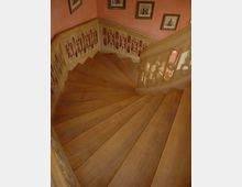 Holztreppe in einem Innenraum mit geschwungenem Verlauf, dekorativ geschnitztem Geländer und rosafarben gestrichenen Wänden. An den Wänden hängen mehrere gerahmte Bilder.