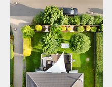 Luftaufnahme eines Einfamilienhauses mit gepflegtem Garten. Der Garten umfasst eine Rasenfläche, Bäume, Büsche, eine Terrasse mit Sonnensegel sowie angrenzenden Gehweg und Straße, an der ein Auto parkt.