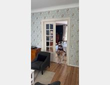 Wohnzimmer mit hellblauer Tapete im Blumenmuster, Holzboden und einem grauen Sessel im Vordergrund. Im Türdurchgang mit verglasten weißen Flügeltüren ist ein Esstisch sichtbar, sowie ein Hund, der im Türrahmen steht.