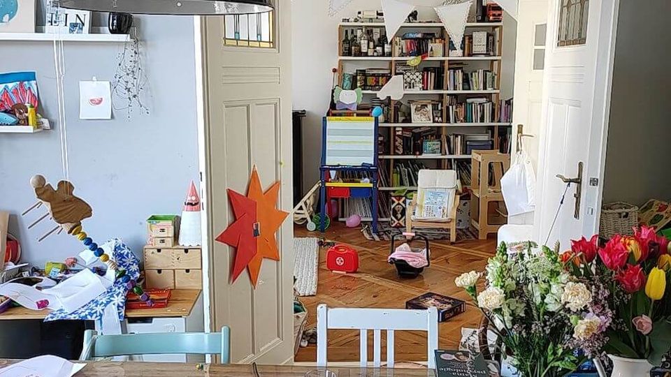 A cluttered home workspace with a wooden table holding a laptop, flowers, books, and various items. In the background, double white doors open into a playroom or study with bookshelves, toys, and colorful decorations, including hanging bunting.