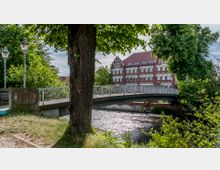 Eine Brücke über einen fließenden Fluss, umgeben von Bäumen und Laternen. Im Hintergrund ist ein großes Backsteingebäude mit weißen Fensterrahmen und Zierelementen zu sehen.