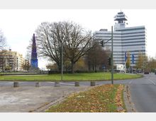 Straßenszene mit einem modernen Bürogebäude im Hintergrund, dem rbb-Fernsehzentrum, und einer Grünfläche mit kahlen Bäumen. Im Vordergrund sind Verkehrswege, Gehwege und eine blaue spitze Skulptur zu sehen, flankiert von Straßenlaternen und Ampeln.