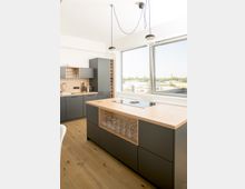 Moderne Küche mit grauen Schränken, einer Insel mit Holzarbeitsplatte und einem integrierten Gläserregal. Große Fenster lassen viel Tageslicht herein und bieten Blick auf eine städtische Landschaft.