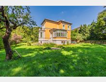 Freistehende, zweigeschossige Villa mit gelb-orangefarbener Fassade, grauem Sockel und verglastem Erker, umgeben von einem großen, grünen Garten. Im Vordergrund liegt eine Rasenfläche mit altem Baum; im Hintergrund stehen dichtes Buschwerk und hohe Bäume unter blauem Himmel.