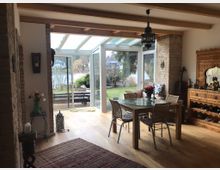 Wohnzimmer mit hellem Holzfußboden, einem Esstisch aus Glas mit vier Stühlen und einer großen Fensterfront, die Blick auf einen Garten und einen See bietet. Rechts steht ein kunstvoll geschnitztes Sideboard mit dekorativen Elementen, während freiliegende Steinwände und eine orientalische Lampe den Raum schmücken.