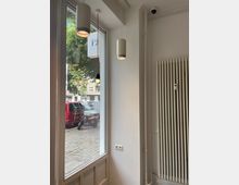 Innenansicht eines Raumes mit großer Fensterscheibe, durch die man eine Straße mit parkenden Fahrzeugen, darunter ein roter Lieferwagen und ein Roller, sieht. An der Decke hängen zwei zylindrische Lampen, rechts steht ein weißer Rippenheizkörper an der Wand.