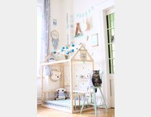 Ein helles Kinderzimmer mit einer kleinen Hausbett-Konstruktion aus Holz, dekoriert mit einer Lichterkette und bunten Kugeln. An der weißen Wand hängen kindgerechte Dekorationen wie sternförmige Ornamente, Regale und Stofftiere; ein gestreifter Stuhl mit einer sitzenden Katze steht neben dem Bett.