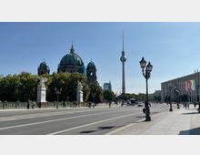 Straßenansicht in Berlin mit Blick auf den Berliner Dom, dem Fernsehturm und einer Brücke mit Statuen und Laternen. Im Hintergrund sind Bürogebäude und klare Wetterverhältnisse mit blauem Himmel zu sehen.