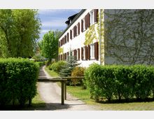 Ein weißes Gebäude mit braunen Fensterläden, teils von Kletterpflanzen bewachsen, steht neben einem schmalen Gehweg, der durch eine grüne Gartenlandschaft mit Büschen, Bäumen und gepflegten Rasenflächen führt. Im Vordergrund befindet sich eine Holzabsperrung.